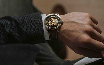 Nahaufnahme einer männlichen Hand mit einer eleganten Armbanduhr mit sichtbar mechanischem Uhrwerk am Handgelenk, getragen zu dunkler Kleidung.