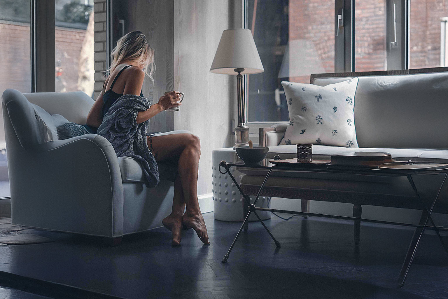 Eine Frau sitzt in einem hellen Wohnzimmer in einem Sessel nahe eines großen Fensters. Sie hält eine Tasse in der Hand und hat eine Decke locker um ihre Schultern gelegt. Vor ihr steht ein Couchtisch mit Schalen, Büchern und Dekoration, im Hintergrund ein Sofa mit Kissen.